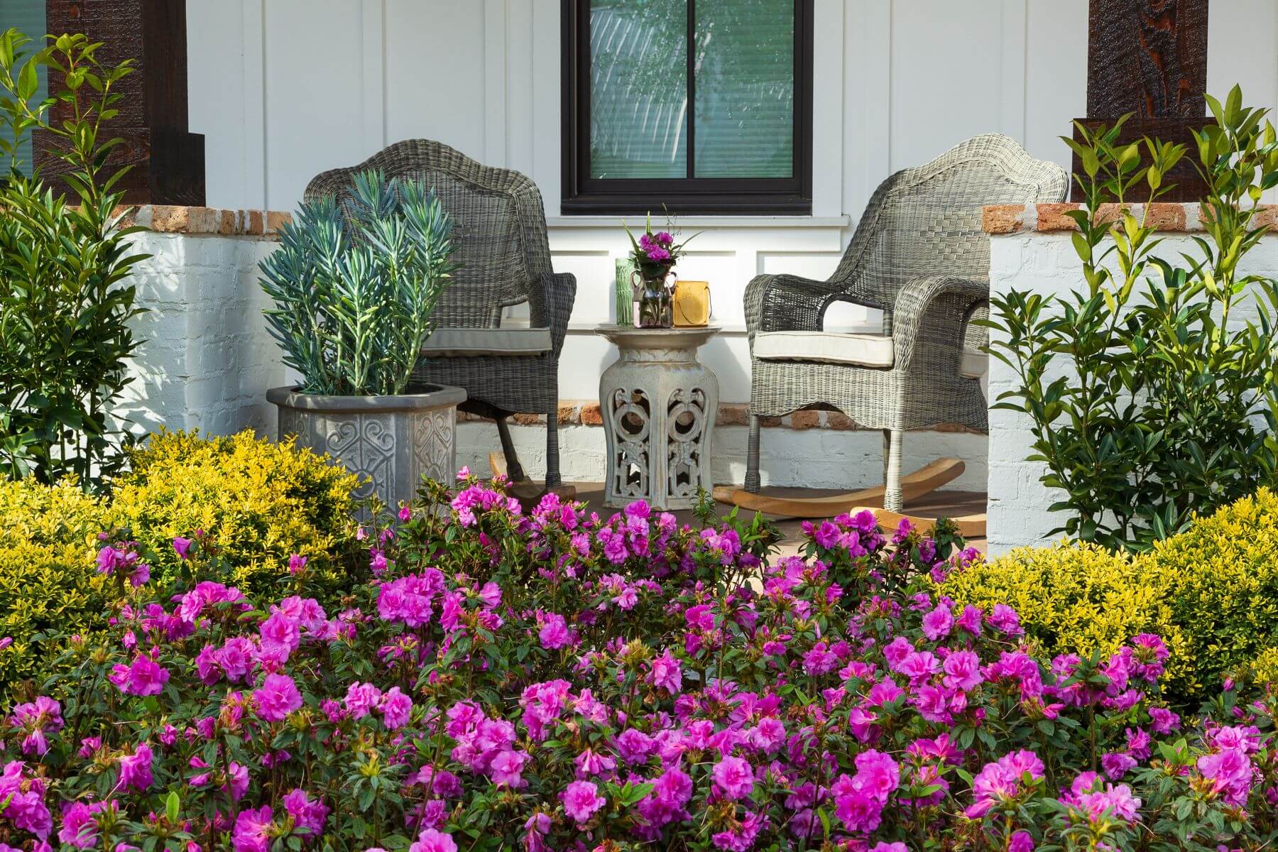 Outdoor seating area with wicker chairs and a small table on a patio, surrounded by flowers and plants featuring Encore Azalea Autumn Majesty with it's purple blooms