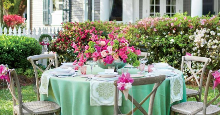 Outdoor garden party setup with a table covered in a light green tablecloth, pink flowers, and four wooden chairs, in front of a white house with blooming bushes.